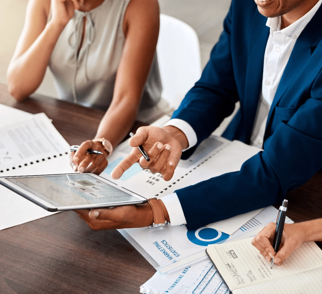 Medium close-up view of financial brokers looking over a tablet and notebooks of financial info at a conference table.